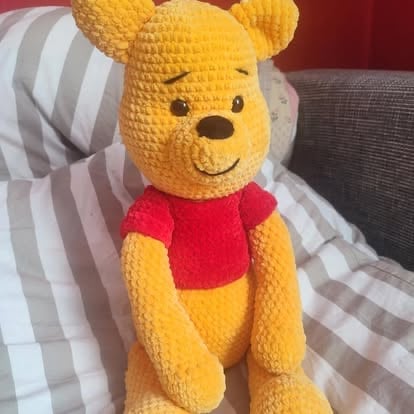Handmade yellow crocheted Winnie the Pooh bear toy wearing a red shirt, sitting on striped bedding
