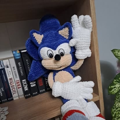 Crocheted Sonic the Hedgehog plush toy in blue with white gloves on a wooden shelf next to books