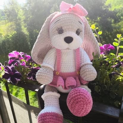 Crocheted pink and white bunny rabbit doll with bow sitting on railing with garden flowers in background
