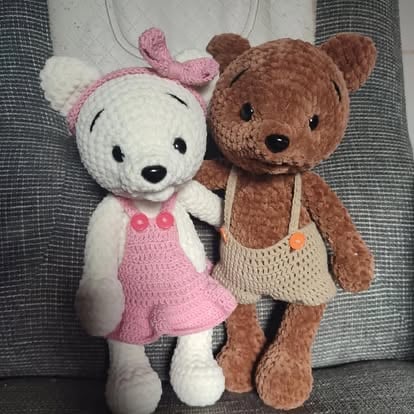 Two handmade crochet teddy bears sitting together on a gray fabric background. One white bear wears a pink dress, the other brown bear wears a cream-colored top