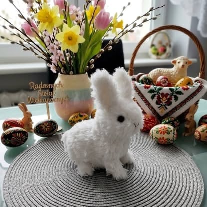 White plush rabbit figurine on circular mat surrounded by Easter decorations including yellow and pink flowers, decorated eggs, and a woven basket
