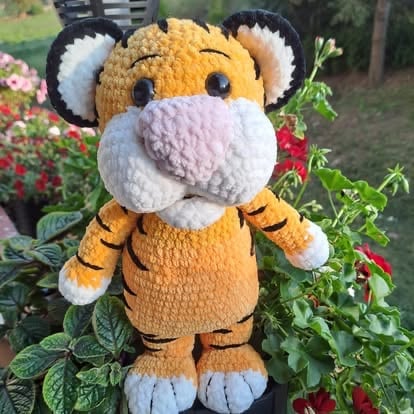 Plush tiger toy with orange and white stripes standing in a garden with red flowers and green foliage in background