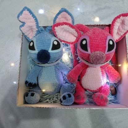 Two plush toys of Stitch characters in blue and pink sitting together with fairy lights