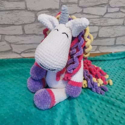 Colorful crocheted unicorn toy with white face, purple mane, and multicolored wavy hair sitting on green mat