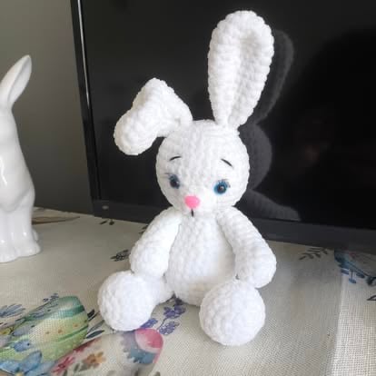 White knitted bunny rabbit plush toy with blue eyes and pink nose sitting on a desk next to a computer monitor